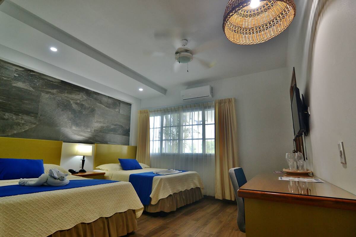 Habitación de hotel con dos camas, un escritorio, una silla, un ventanal con cortinas, un ventilador de techo y un televisor montado en la pared.