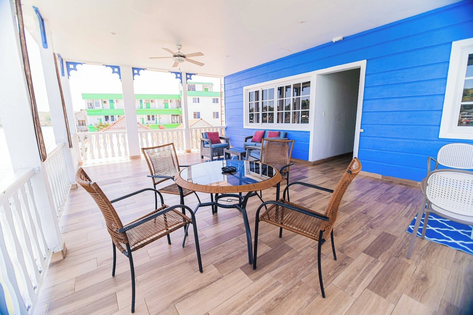 Une terrasse couverte avec une table ronde et des chaises en osier, un ventilateur de plafond et une vue sur un bâtiment doté de toits végétalisés.
