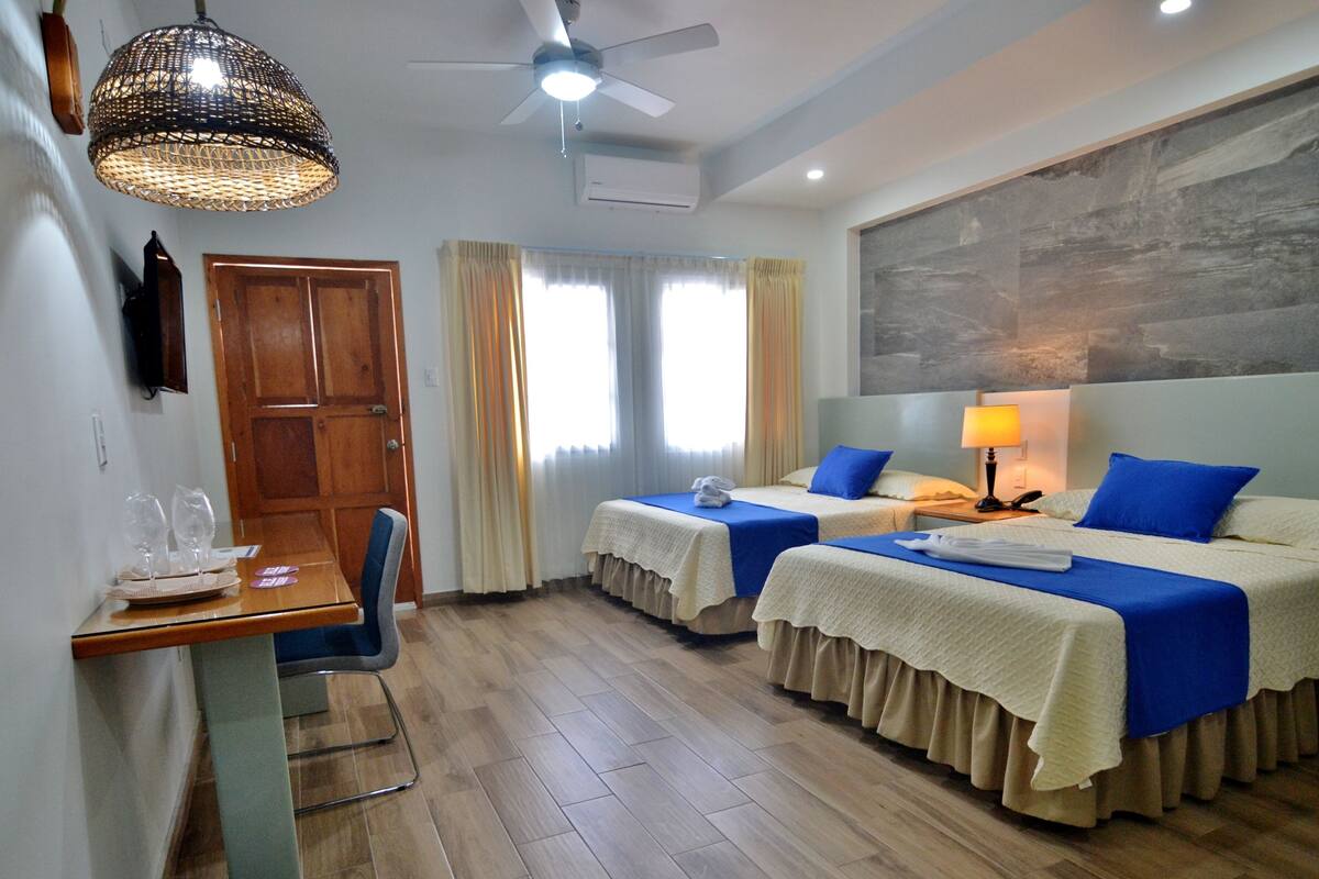 Habitación de hotel con dos camas, un escritorio con silla, un ventilador de techo y un cuadro abstracto grande en la pared.