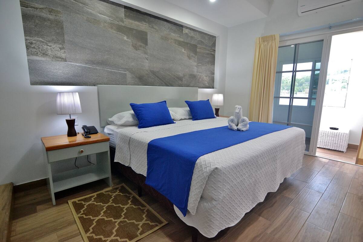 Habitación de hotel ordenada con una cama grande, una mesita de noche, una lámpara y vistas al exterior.
