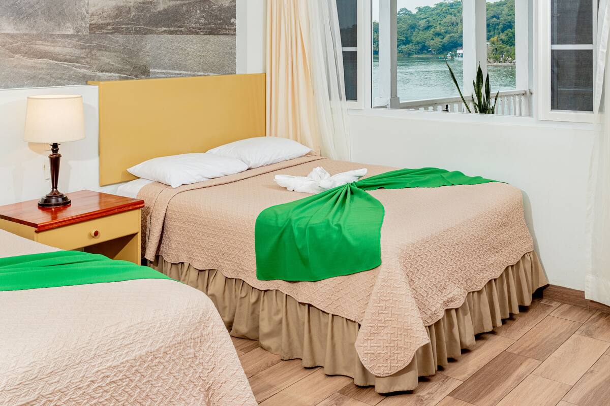 Una habitación de hotel con cama, mesita de noche, lámpara y ventana con vista al lago.