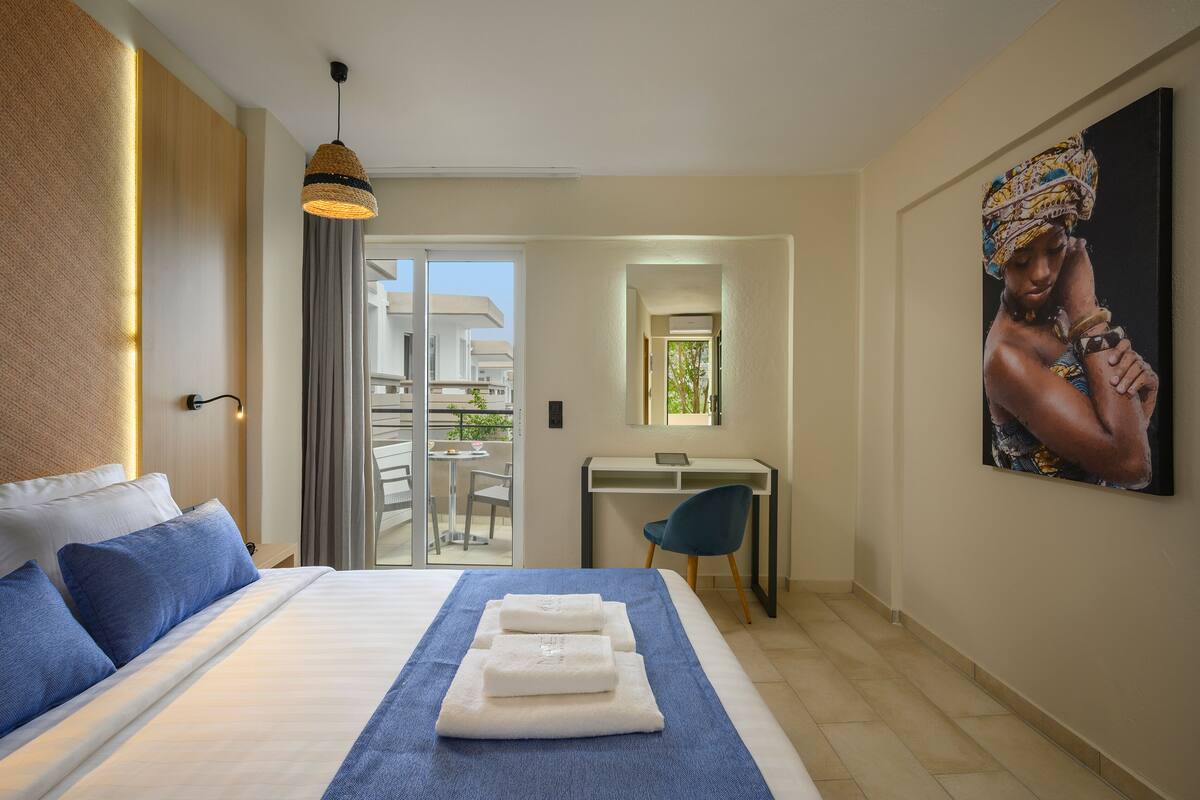 A hotel room with a bed, a desk, a chair, a painting, and a view of the outside.