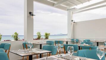 A restaurant with tables and chairs, a view of the sea, and a white ceiling.