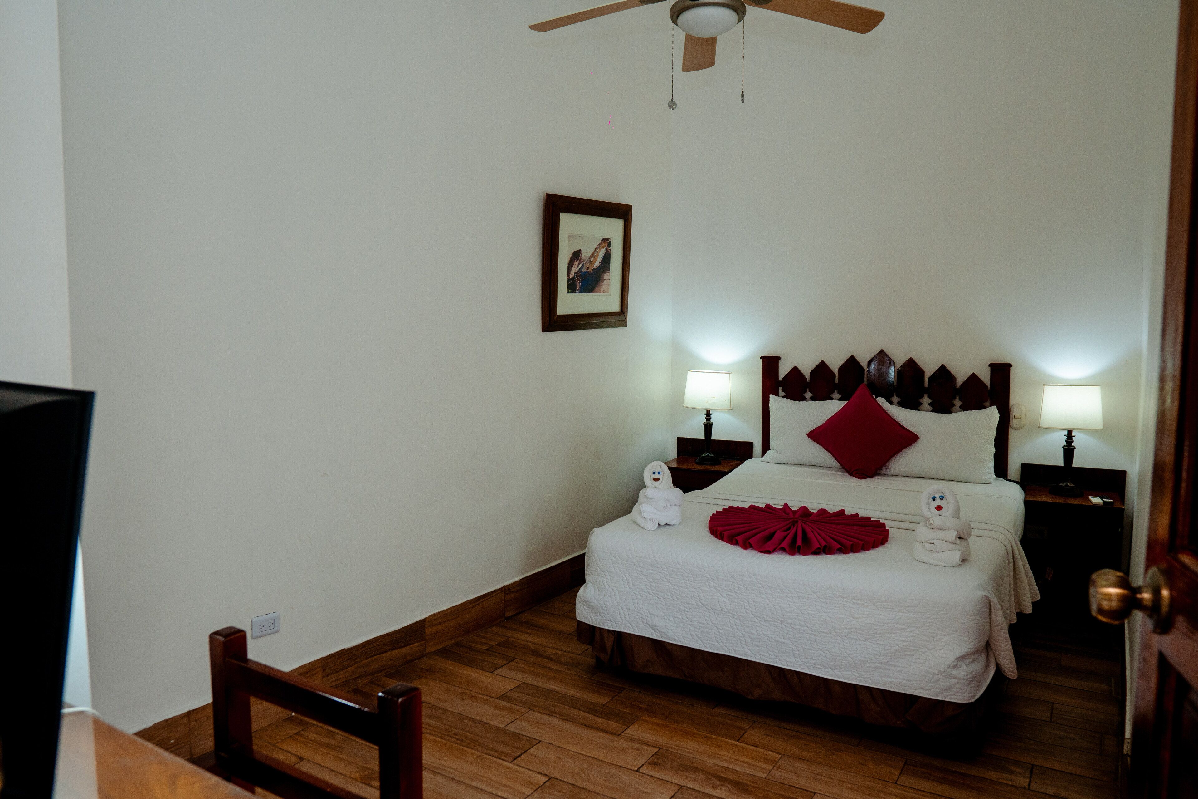 Ein Schlafzimmer mit einem Bett, zwei Nachttischen, einem Deckenventilator, einem gerahmten Bild an der Wand und Holzboden.