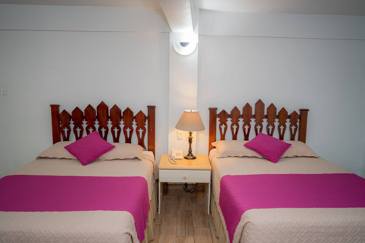 Habitación de hotel con dos camas, cada una con cabecero de madera, mesita de noche con lámpara y teléfono.