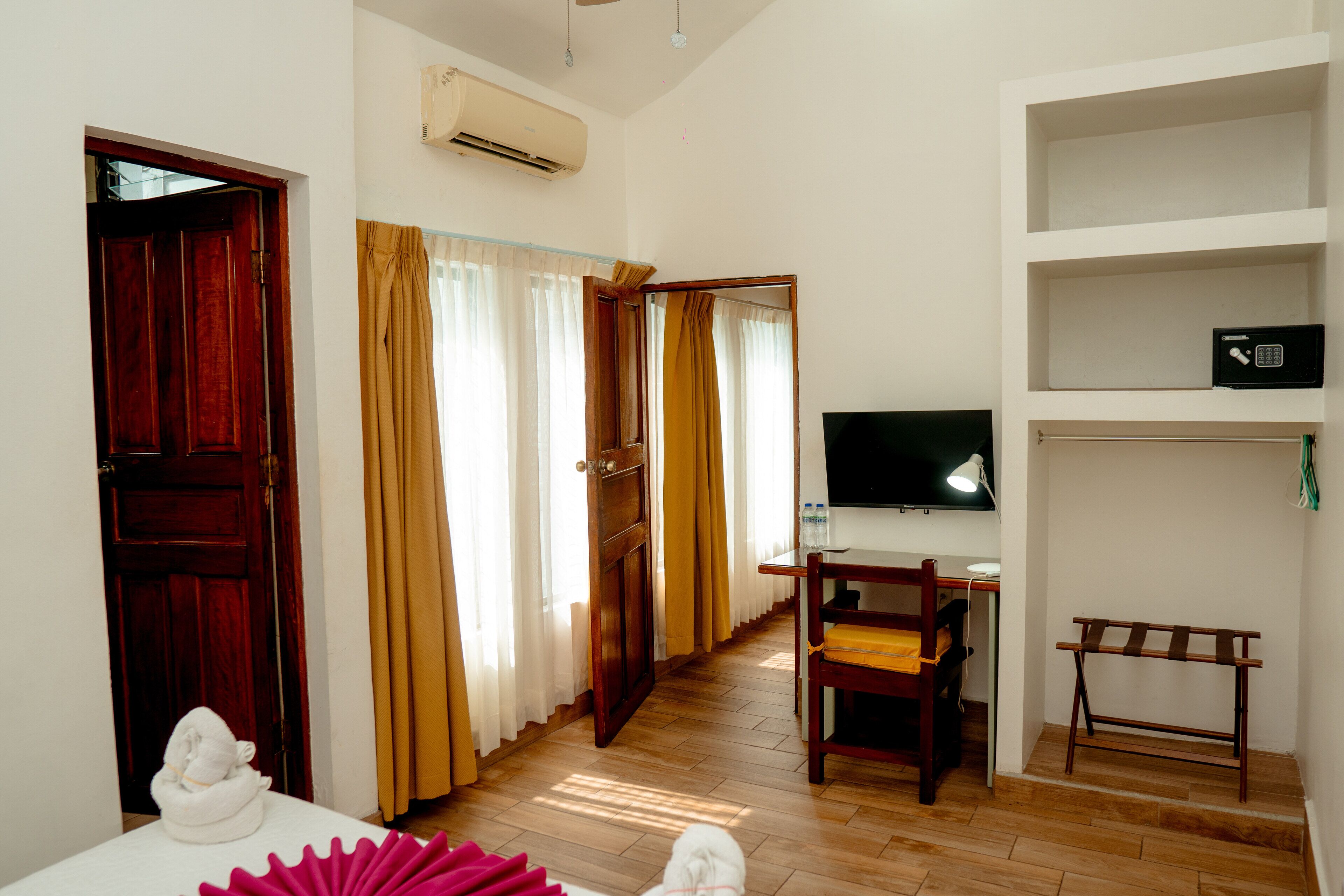 A hotel room with a wooden floor, a desk with a chair, a television, and a shelf with a safe.