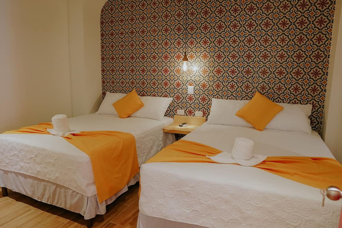 Habitación de hotel con dos camas, cada una con una manta naranja y almohadas blancas. Se observa un papel tapiz decorativo detrás de las camas y una pequeña mesa con una lámpara entre ellas.