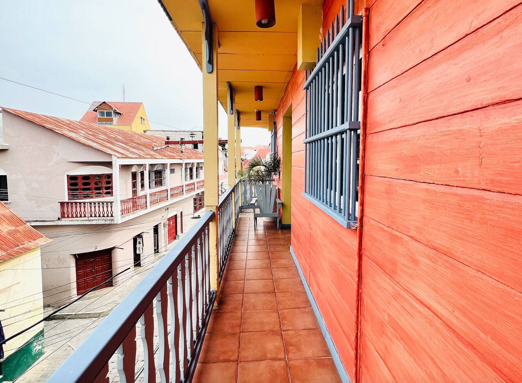 Un balcón estrecho con suelo de baldosas, barandilla y vistas a edificios cercanos.