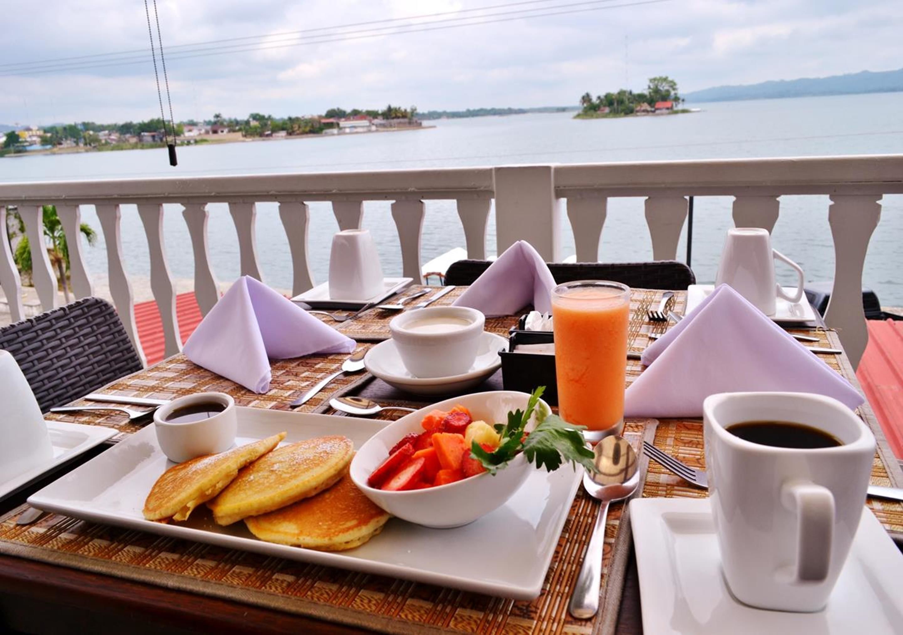 A breakfast table with pancakes, fruit, coffee, and juice, overlooking a lake with distant islands.