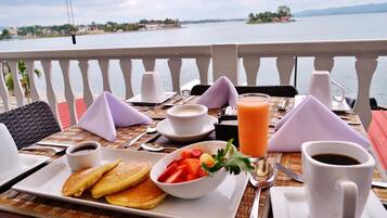 A breakfast table with pancakes, fruit, coffee, and juice, overlooking a lake with distant islands.