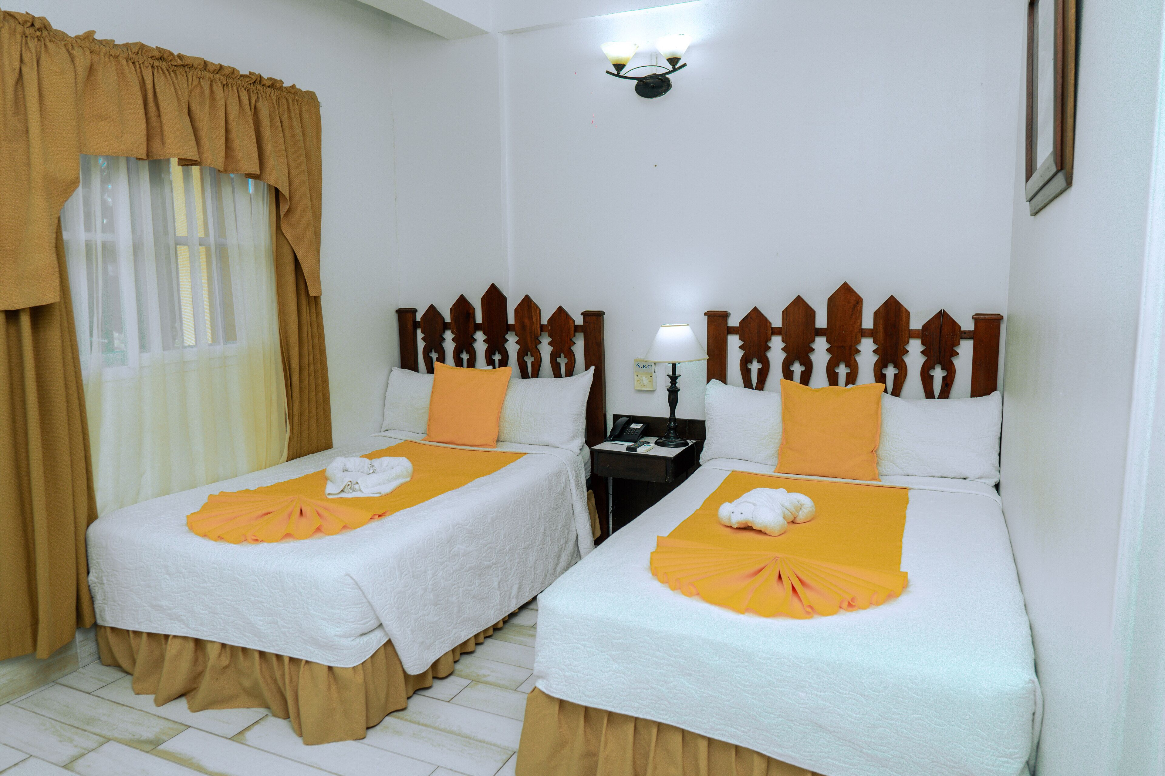 A hotel room with two beds, each with a white bedspread, orange pillows, and a towel on the bed. There is a nightstand with a lamp and a phone.