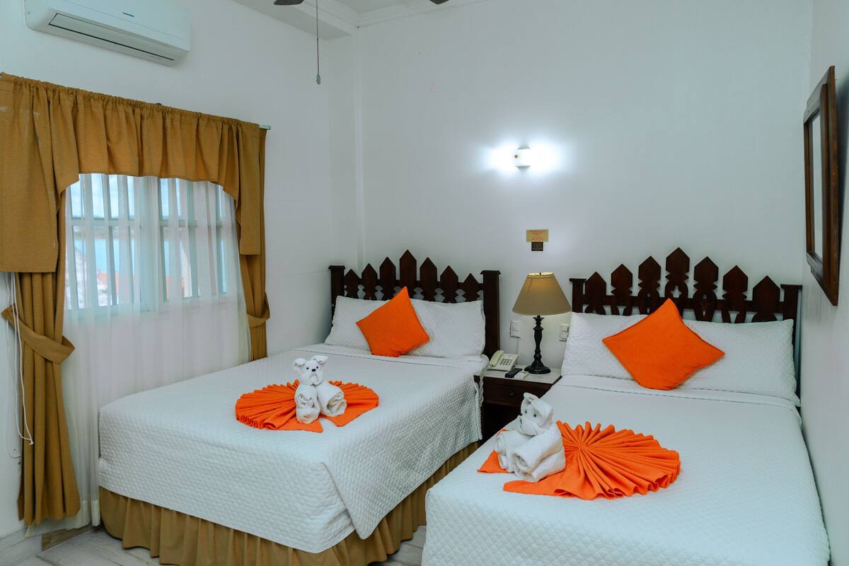 Habitación de hotel con dos camas, cada una con ropa de cama blanca y almohadas naranjas, una mesita de noche con una lámpara, y una ventana con cortinas.
