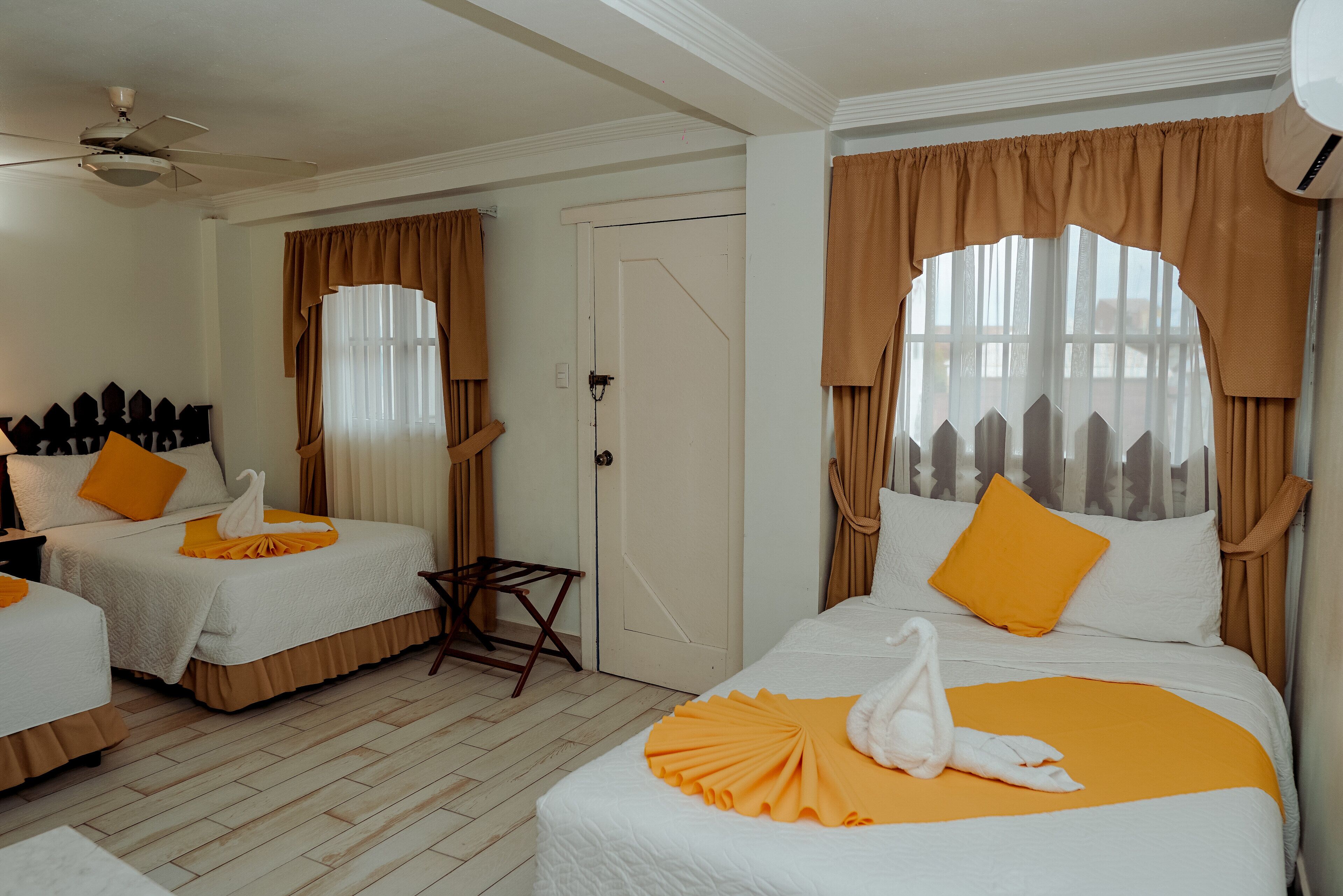 Hotelkamer met twee bedden, elk voorzien van een witte spreidek en oranje accenten, zoals kussens en een plaid. Er is een kleine tafel met een stoel, een plafondventilator en een deur met een raam.