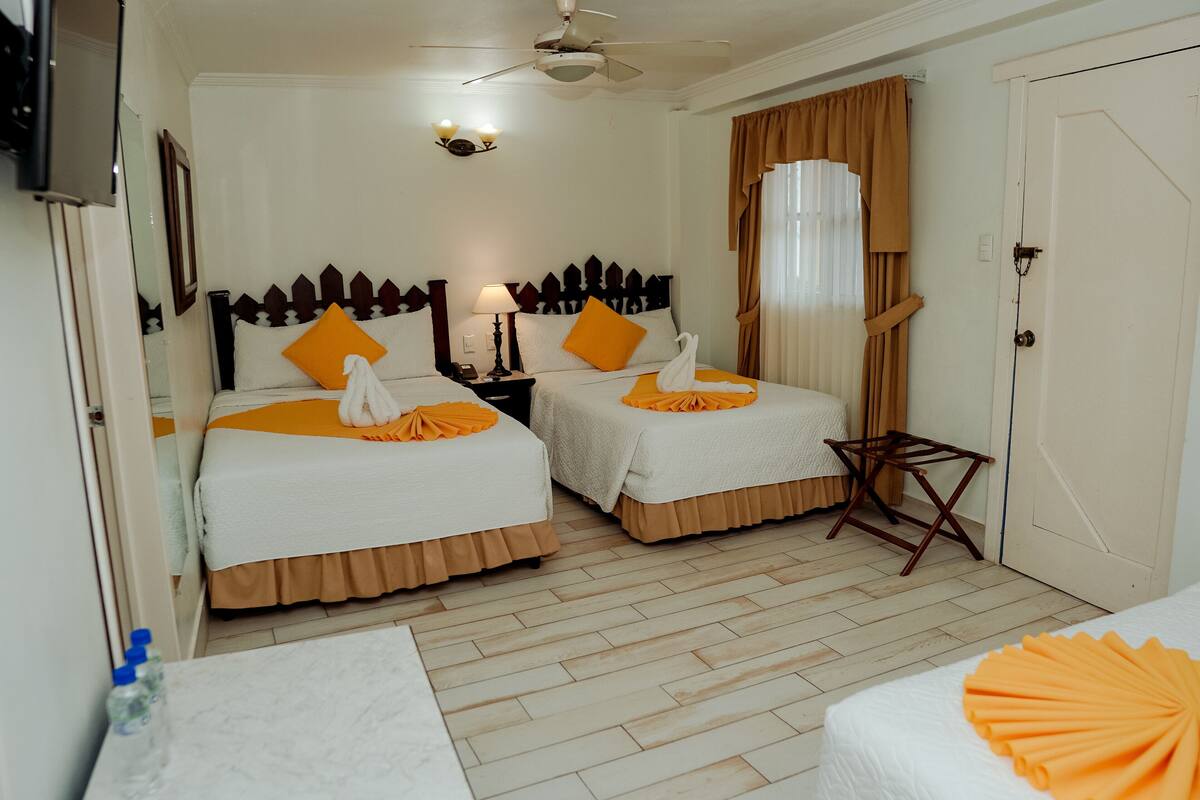 Habitación de hotel con dos camas, un ventilador de techo, una mesita de noche con lámpara, una pequeña mesa y una ventana con cortinas.