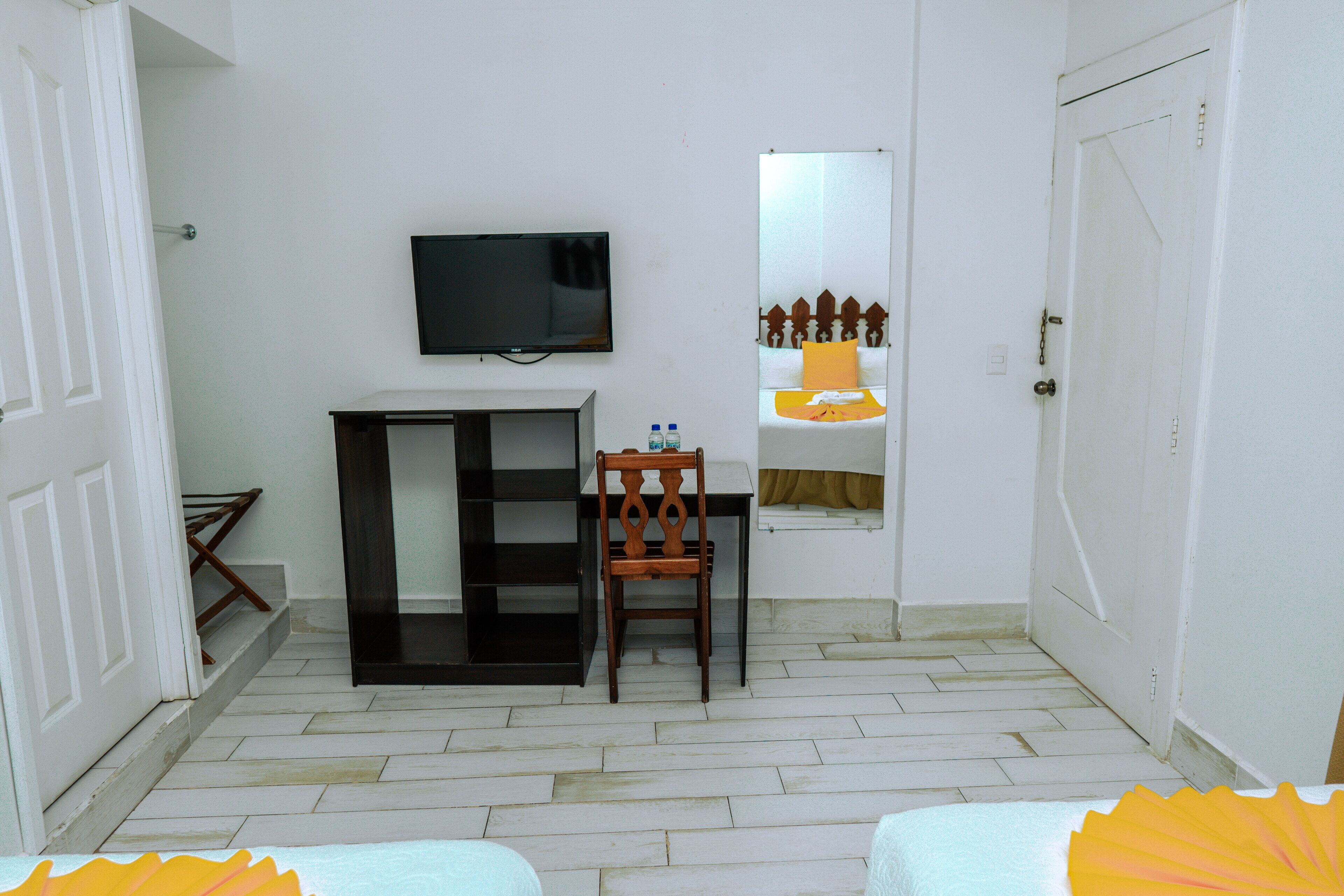 A room with a television mounted on the wall, a wooden chair, a small table, and a mirror reflecting a bed with a yellow pillow.