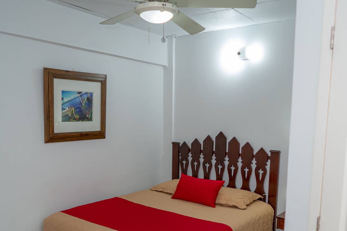 Un dormitorio con una cama de madera, un ventilador de techo, un cuadro en la pared y una puerta blanca.