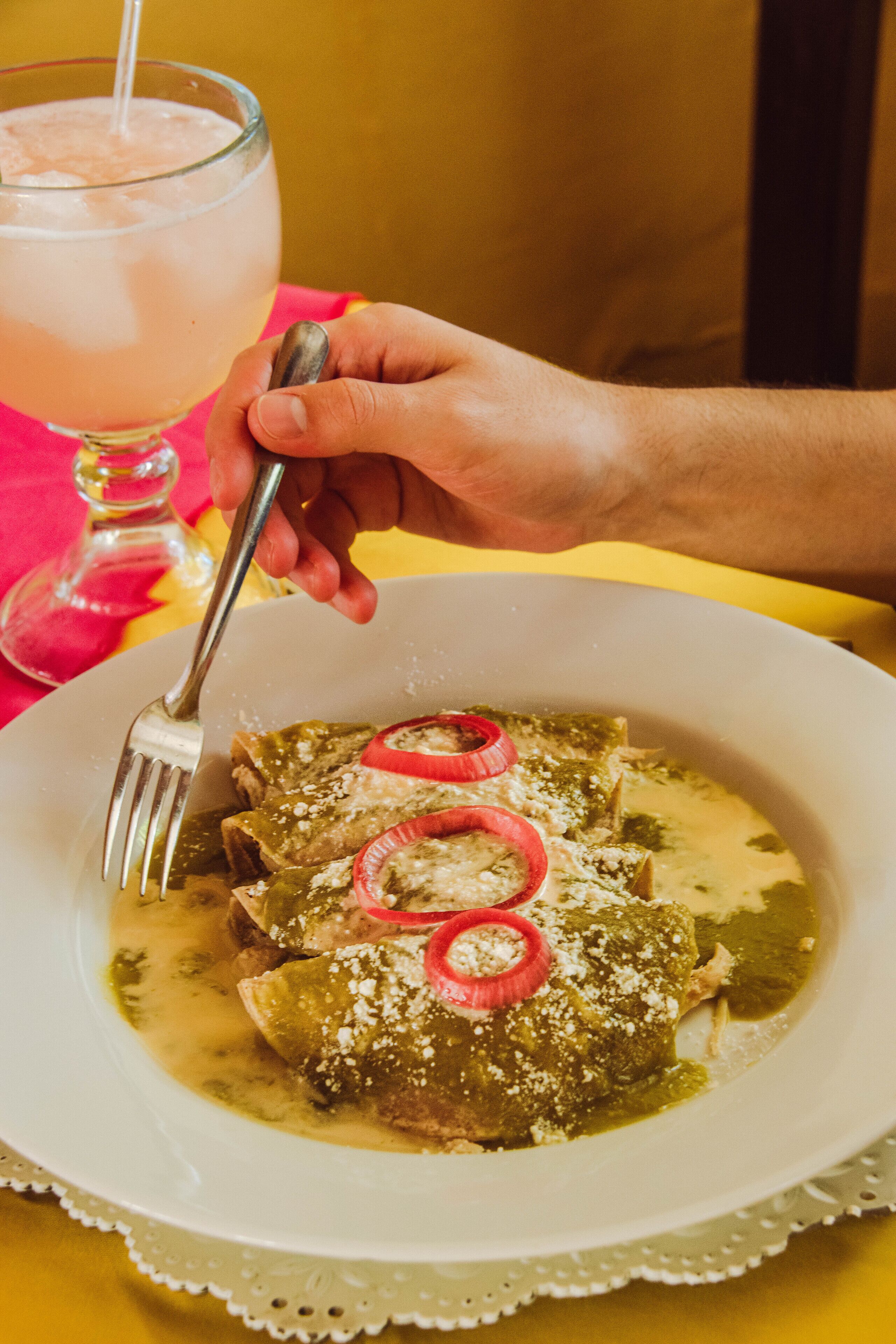 A plate of enchiladas with red onion rings and a glass of pink beverage on a yellow tablecloth.