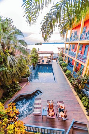 Zona de piscina con sillas de descanso y jacuzzi, rodeada de palmeras y un edificio con balcones.