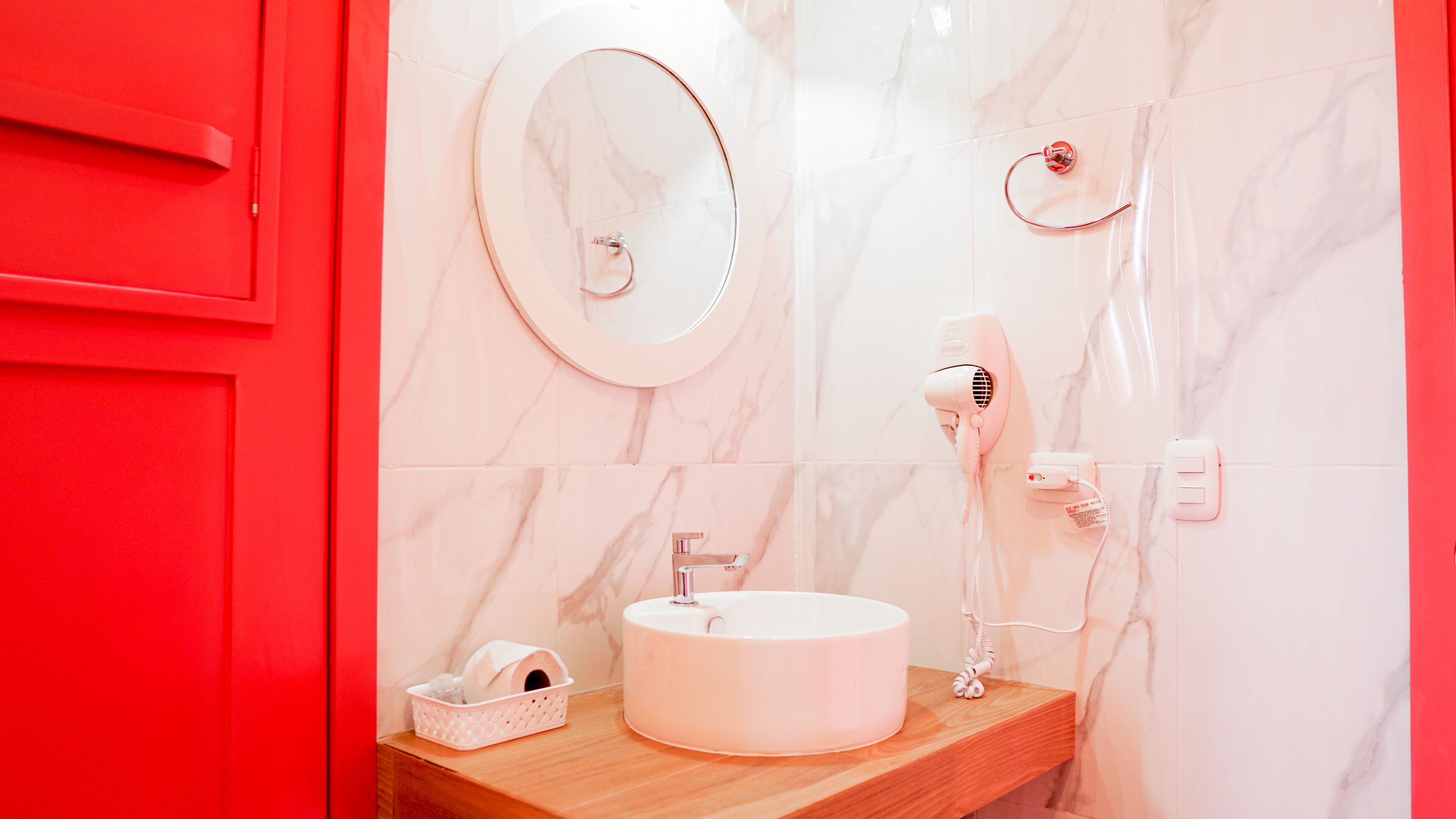 A bathroom with a round mirror, a white sink, a red cabinet, and a hairdryer on the wall.