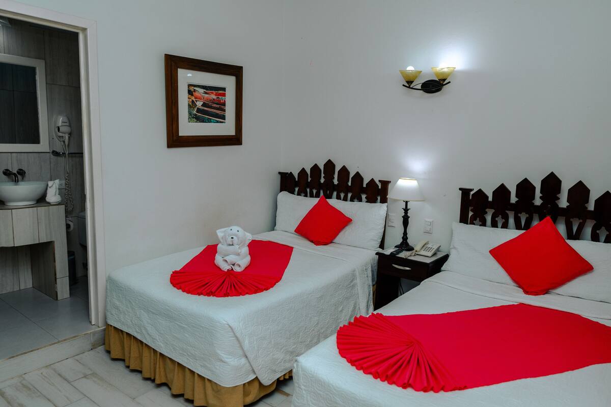 Habitación de hotel con dos camas, cada una con una colcha y almohada rojas, una mesita de noche con lámpara, un cuadro enmarcado en la pared y un baño visible al fondo.