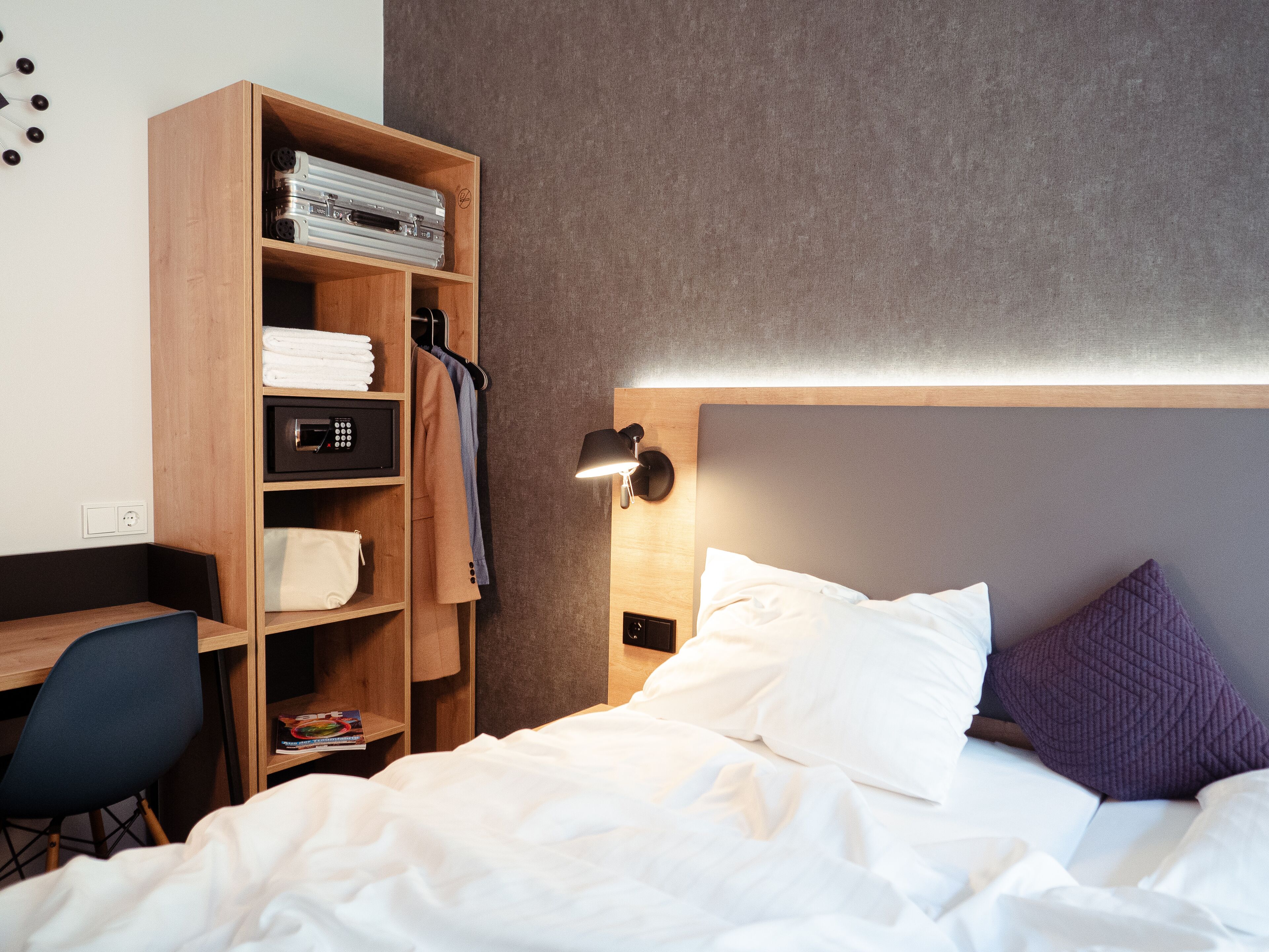 A neatly made bed with white linens, a wooden bedside table with a book, a wall-mounted lamp, and a wardrobe with folded clothes and a suitcase.