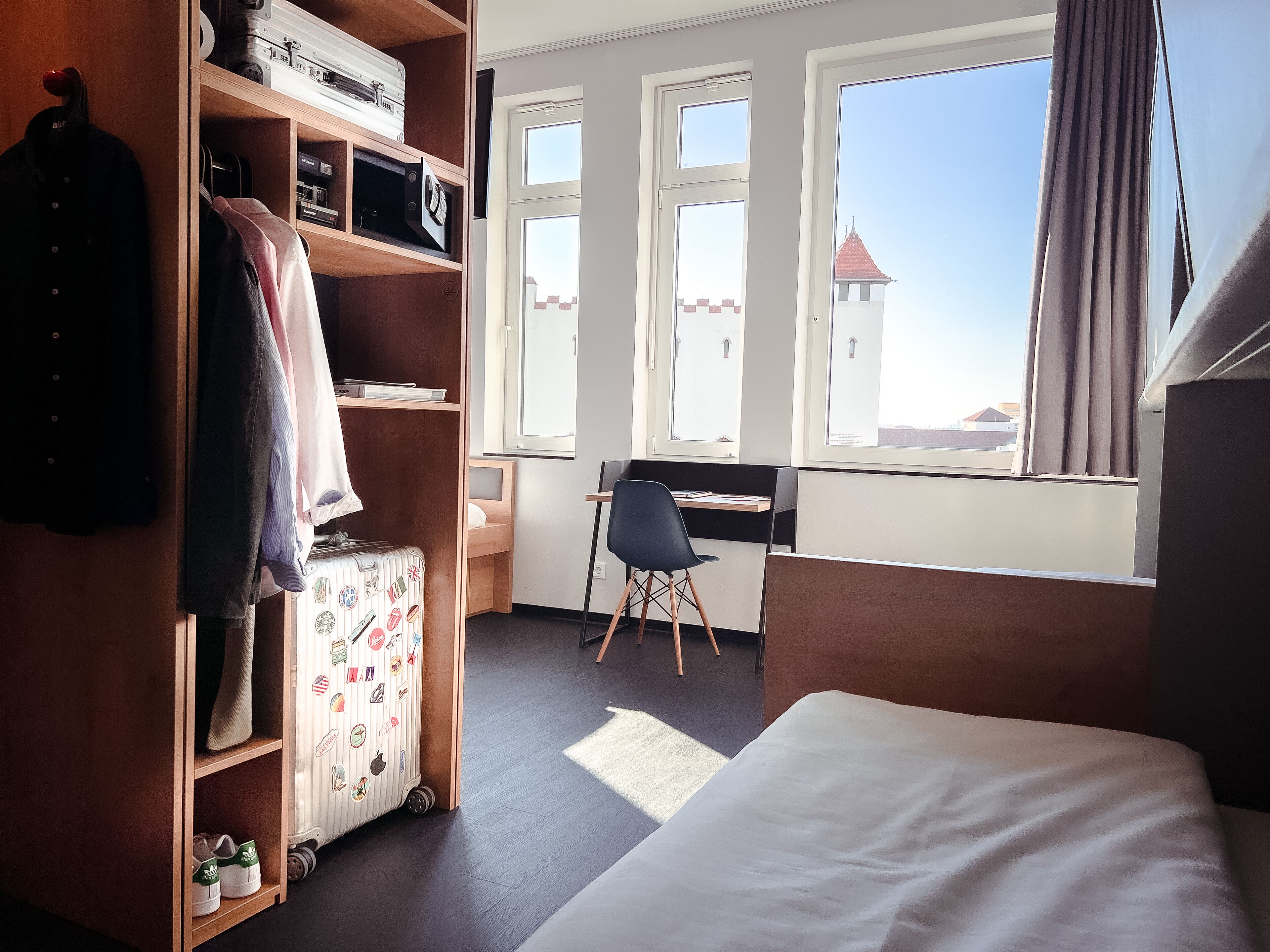 A hotel room with a bed, a desk, a chair, a wardrobe, and a window with a view of buildings.