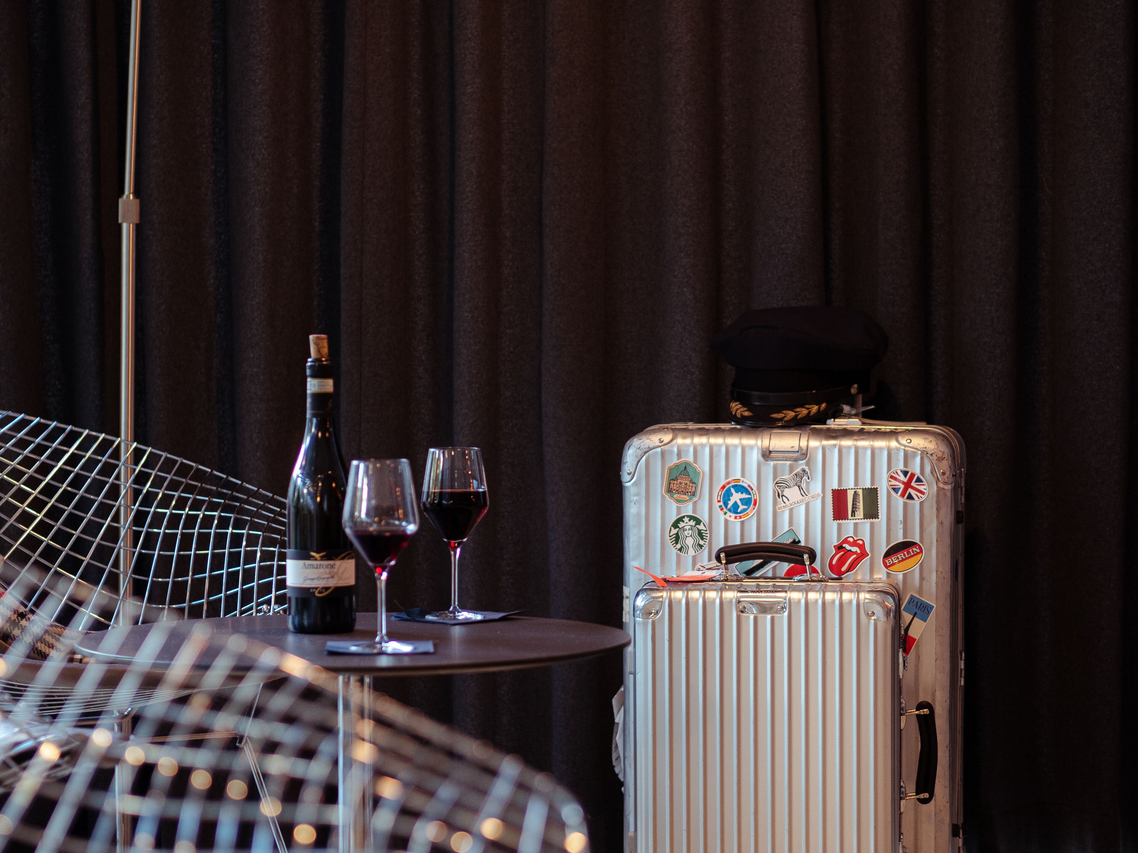 A table with a bottle of wine, two wine glasses, and a suitcase with travel stickers.