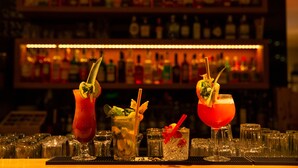 Un bar con varios vasos de cócteles, una variedad de botellas al fondo y una barra con vasos y una caja registradora.