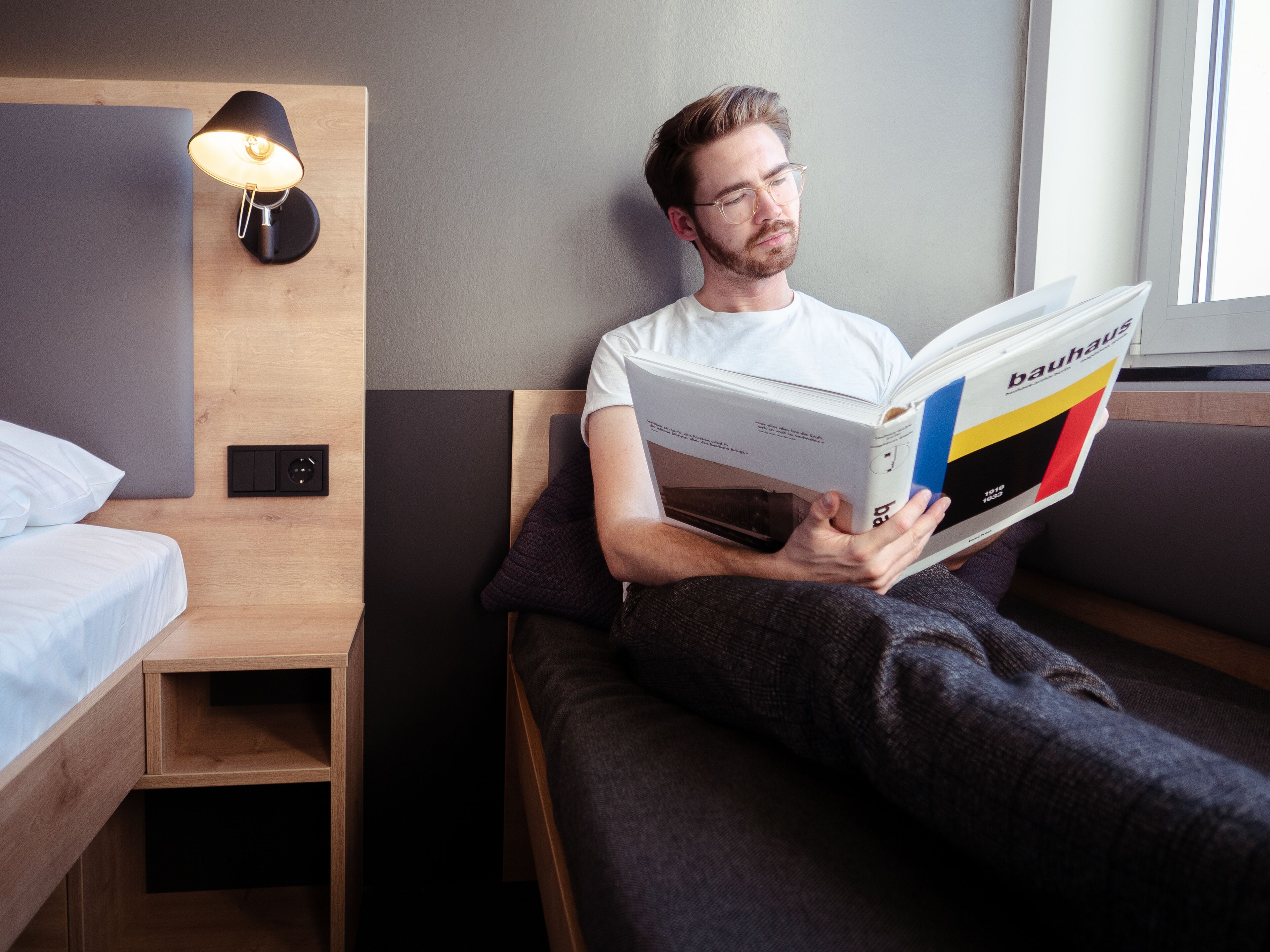 Una persona leyendo un libro en una habitación moderna con una cama, una mesita de noche y una lámpara de pared.