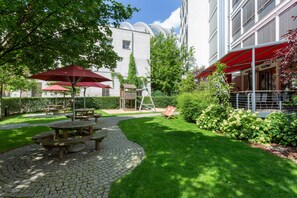 A courtyard with picnic tables and umbrellas, surrounded by greenery and a building with a modern design. - aletto Hotel Kudamm (Berlin)