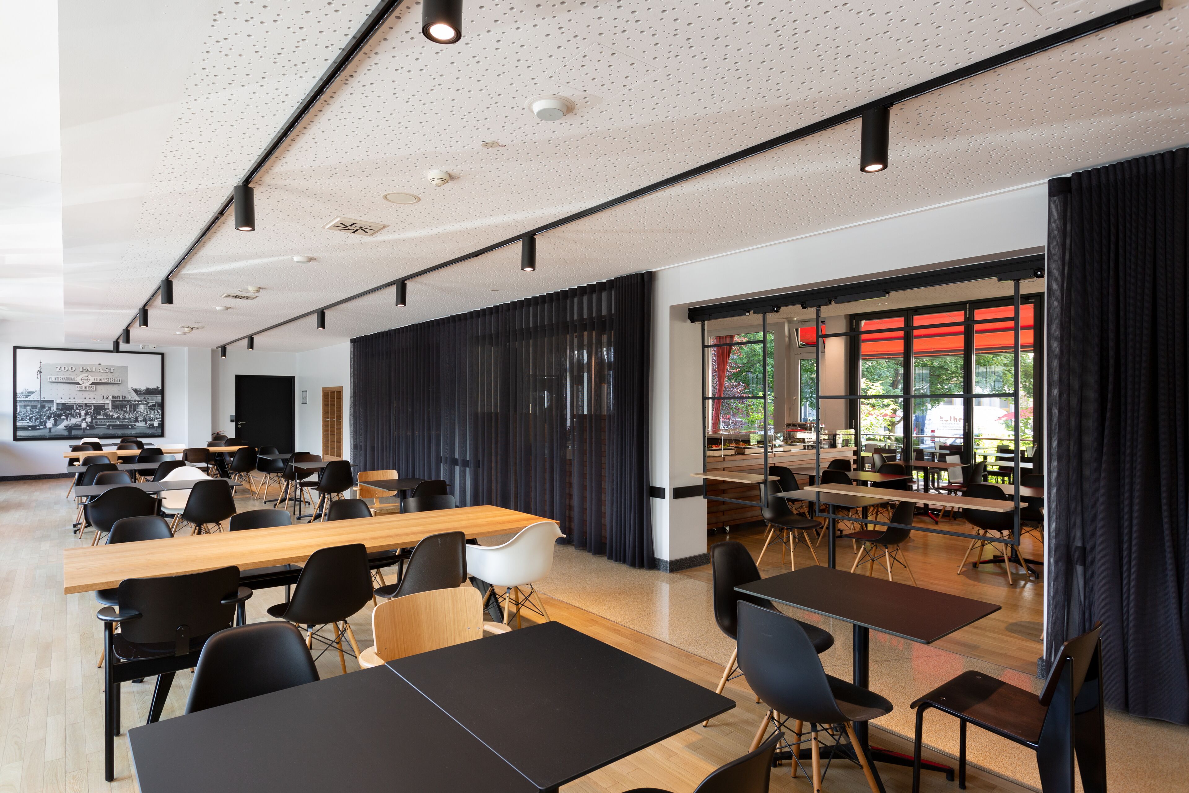 Une salle de conférence moderne, dotée de plusieurs tables et chaises, de grandes fenêtres avec des rideaux rouges et d’un plafond pourvu d’un éclairage encastré.