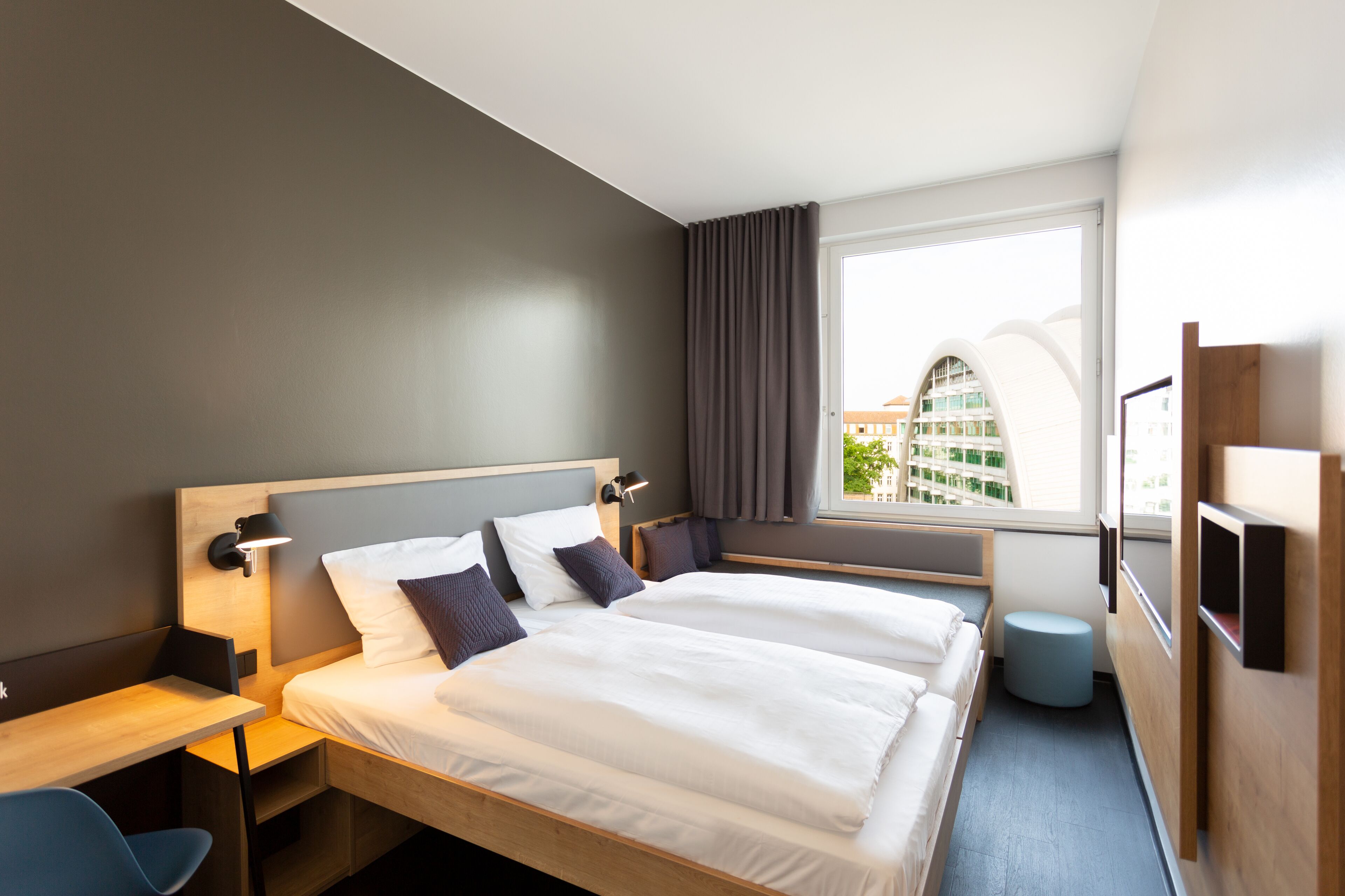 A hotel room with two beds, a desk, a chair, a window with curtains, and a view of buildings outside.