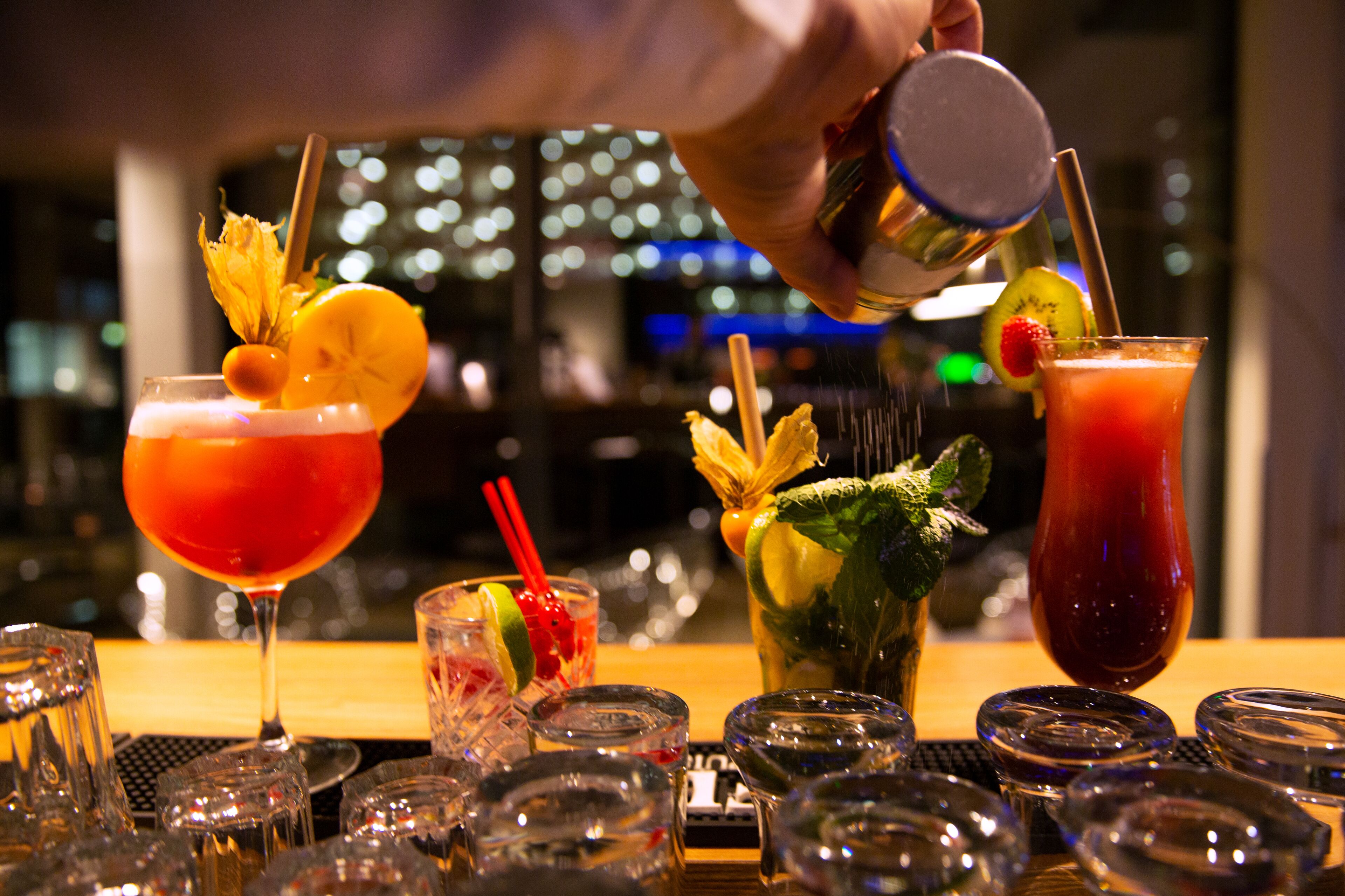 Un bar con diferentes vasos de cócteles, un bartender sirviendo una bebida y un paisaje urbano de fondo.