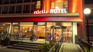 Exterior del hotel Aletto, con un llamativo toldo rojo, entrada de cristal y fachada bien iluminada.