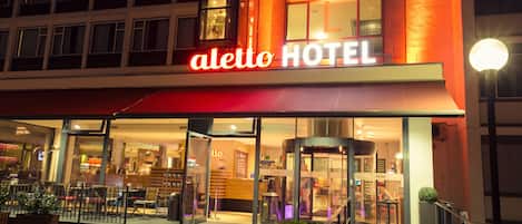 The exterior of the Aletto Hotel with a prominent red awning, glass entrance, and a well-lit facade.