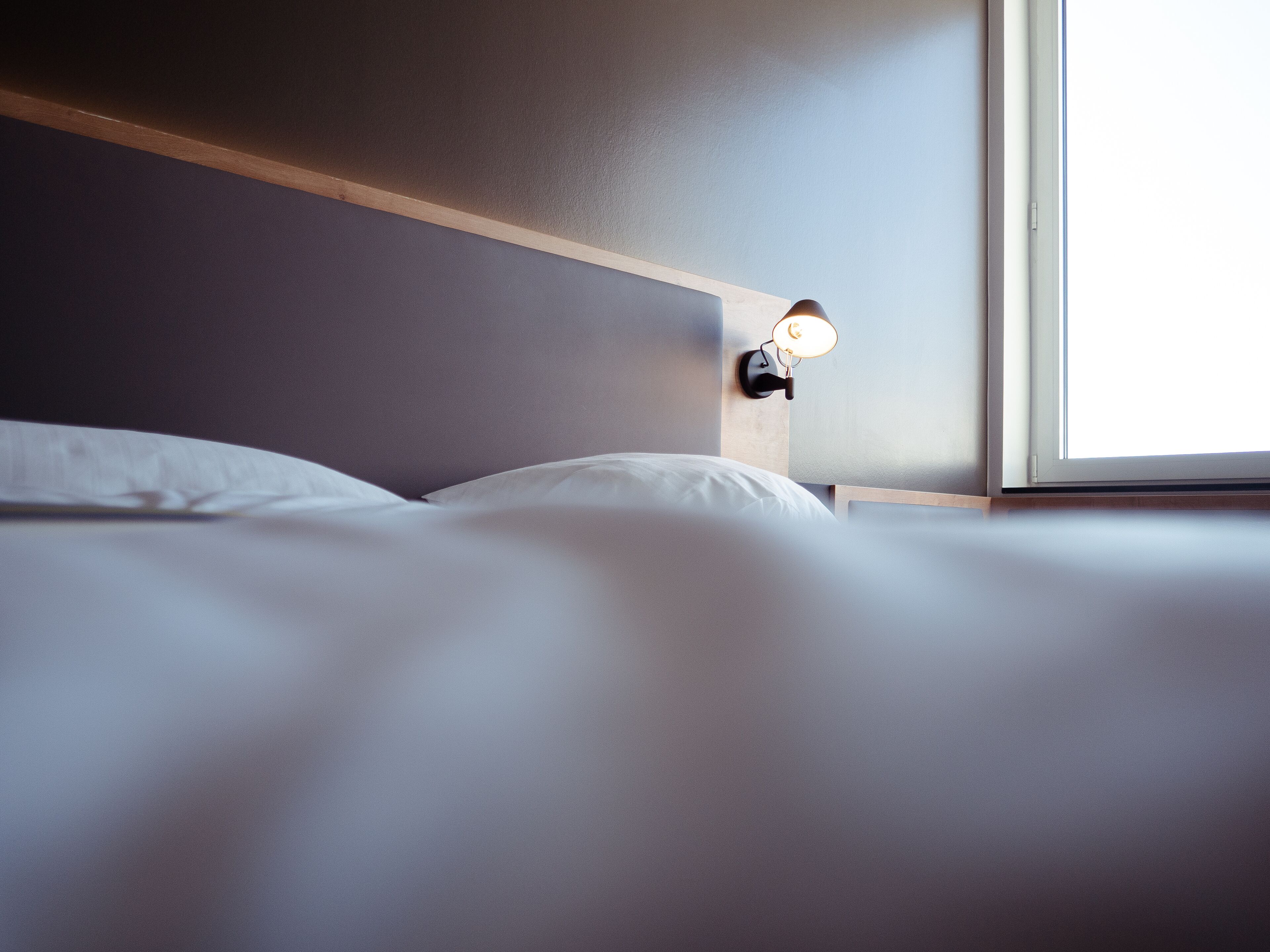 Una cama bien tendida con sábanas blancas, cabecera de madera y lámpara de pared.