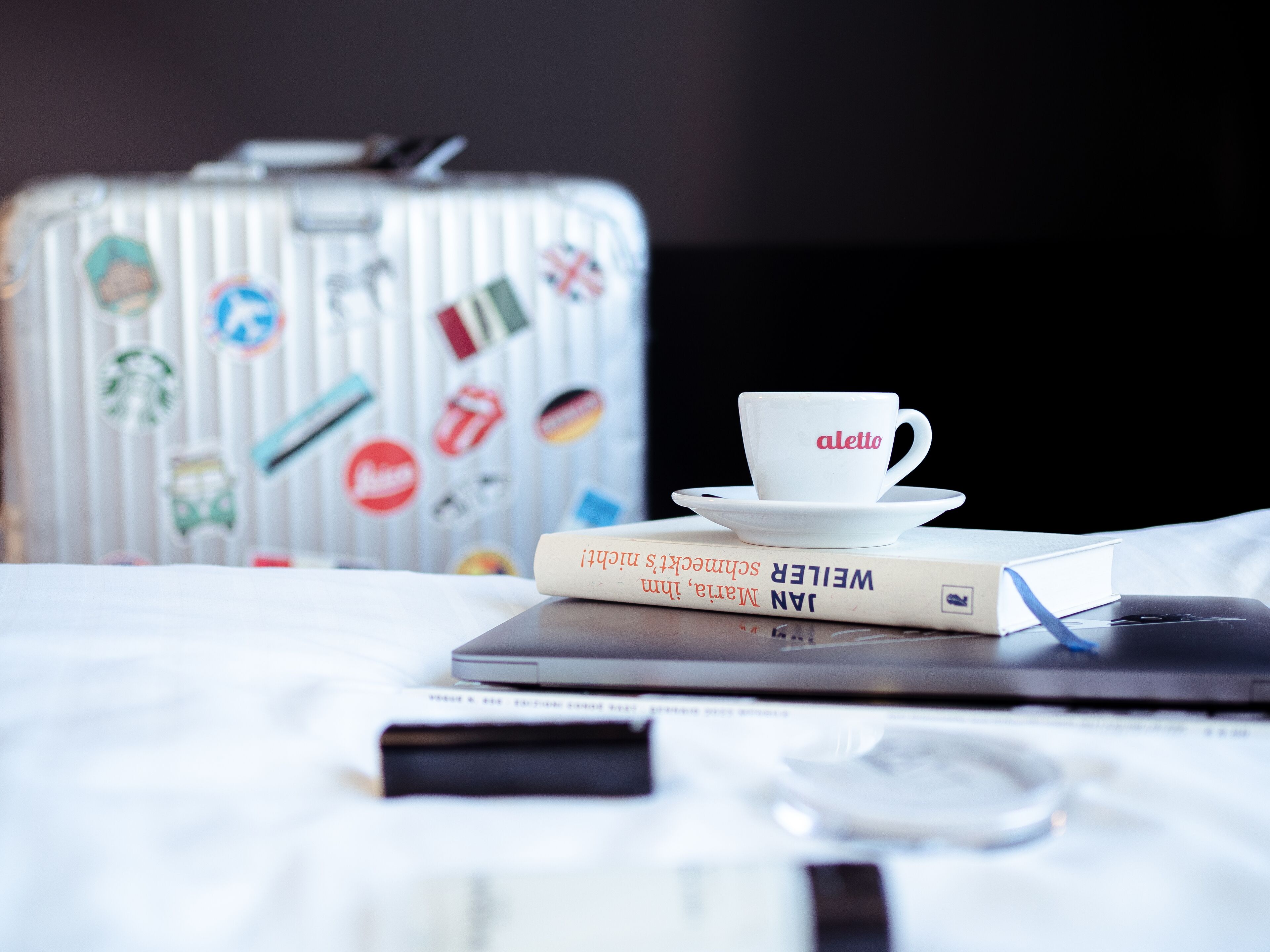 Una cama con una laptop, una taza, un libro y una maleta con calcomanías.