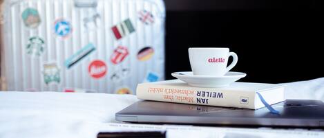Una cama con una laptop, una taza, un libro y una maleta con calcomanías.