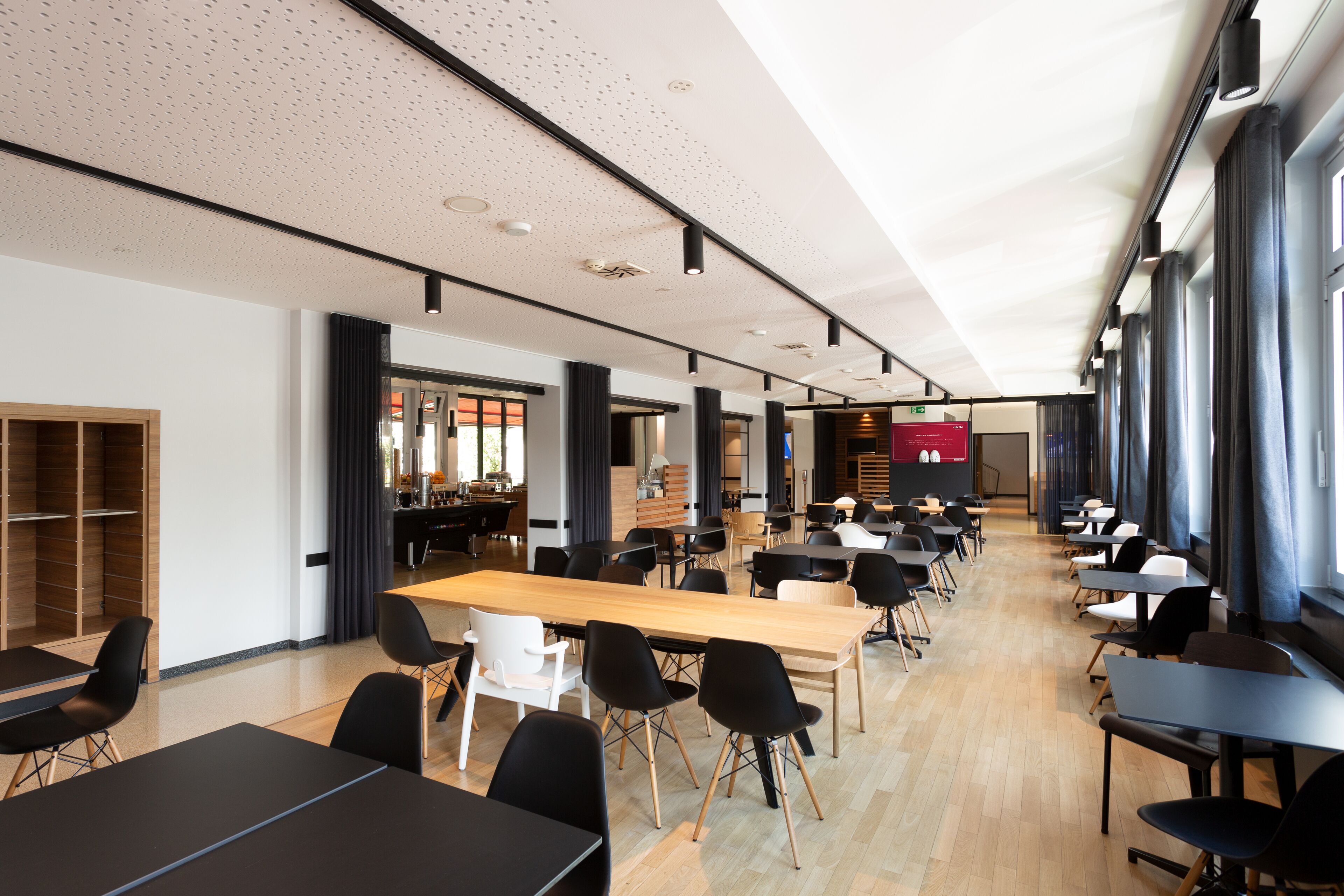 A spacious dining area with long tables and chairs, large windows, and a modern ceiling design.