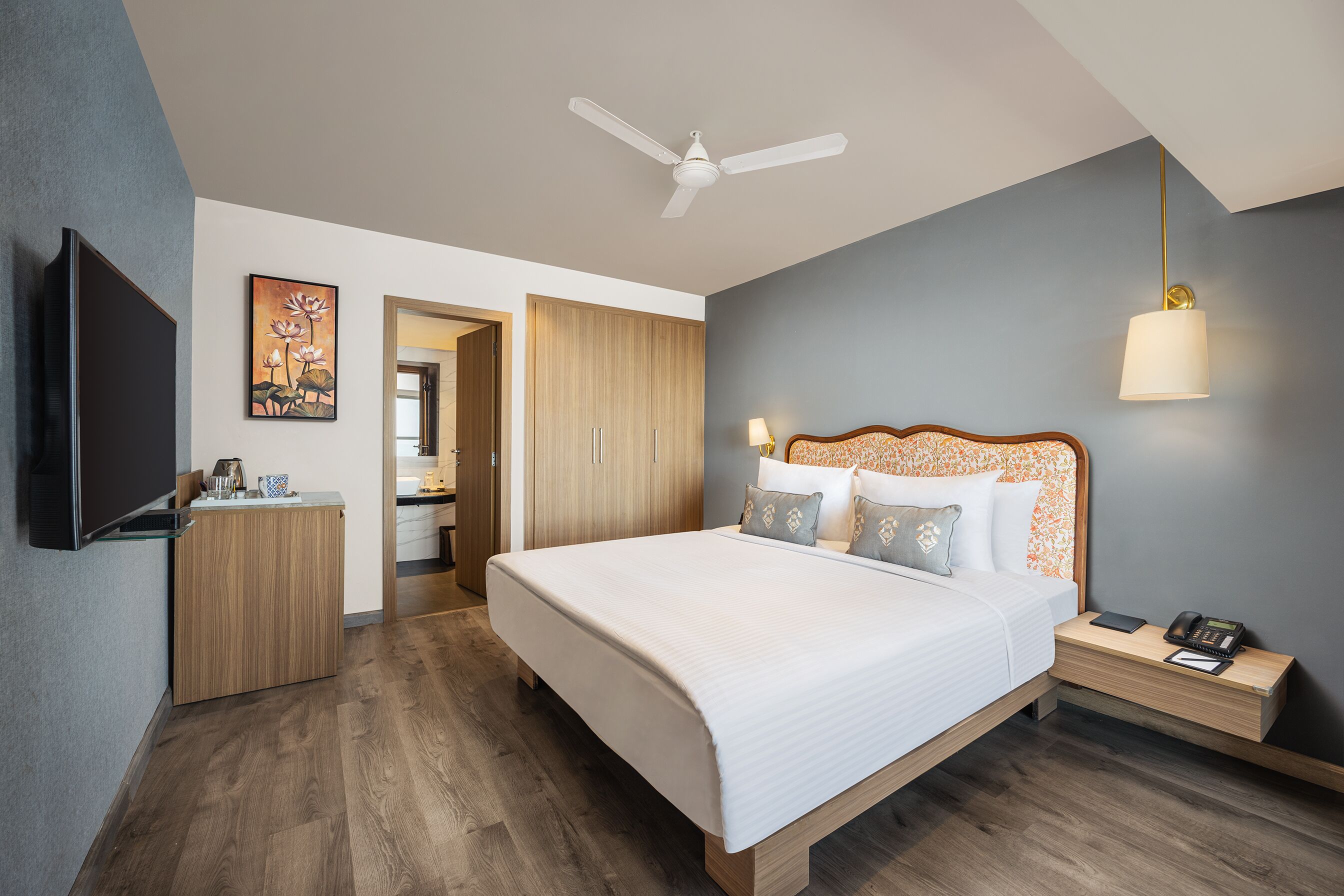 Habitación de hotel moderna con una cama grande, un escritorio con lámpara, un ventilador de techo y un televisor montado en la pared.