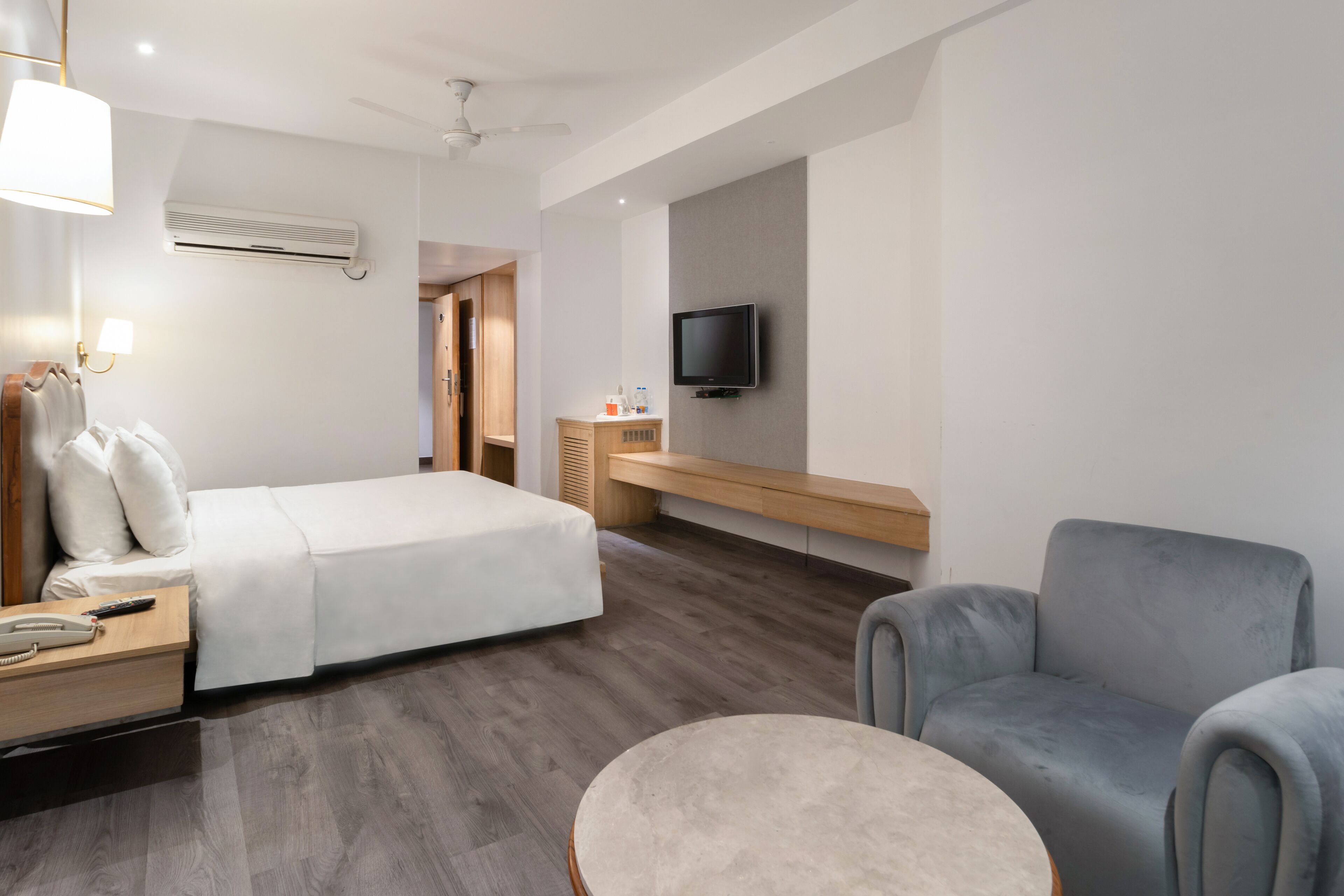 A modern hotel room with a bed, bedside table, lamp, TV, and a grey armchair.