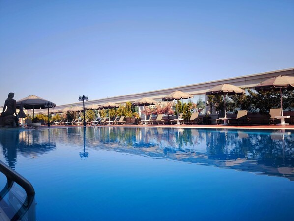 A clear swimming pool with lounge chairs and umbrellas, surrounded by trees and a building.