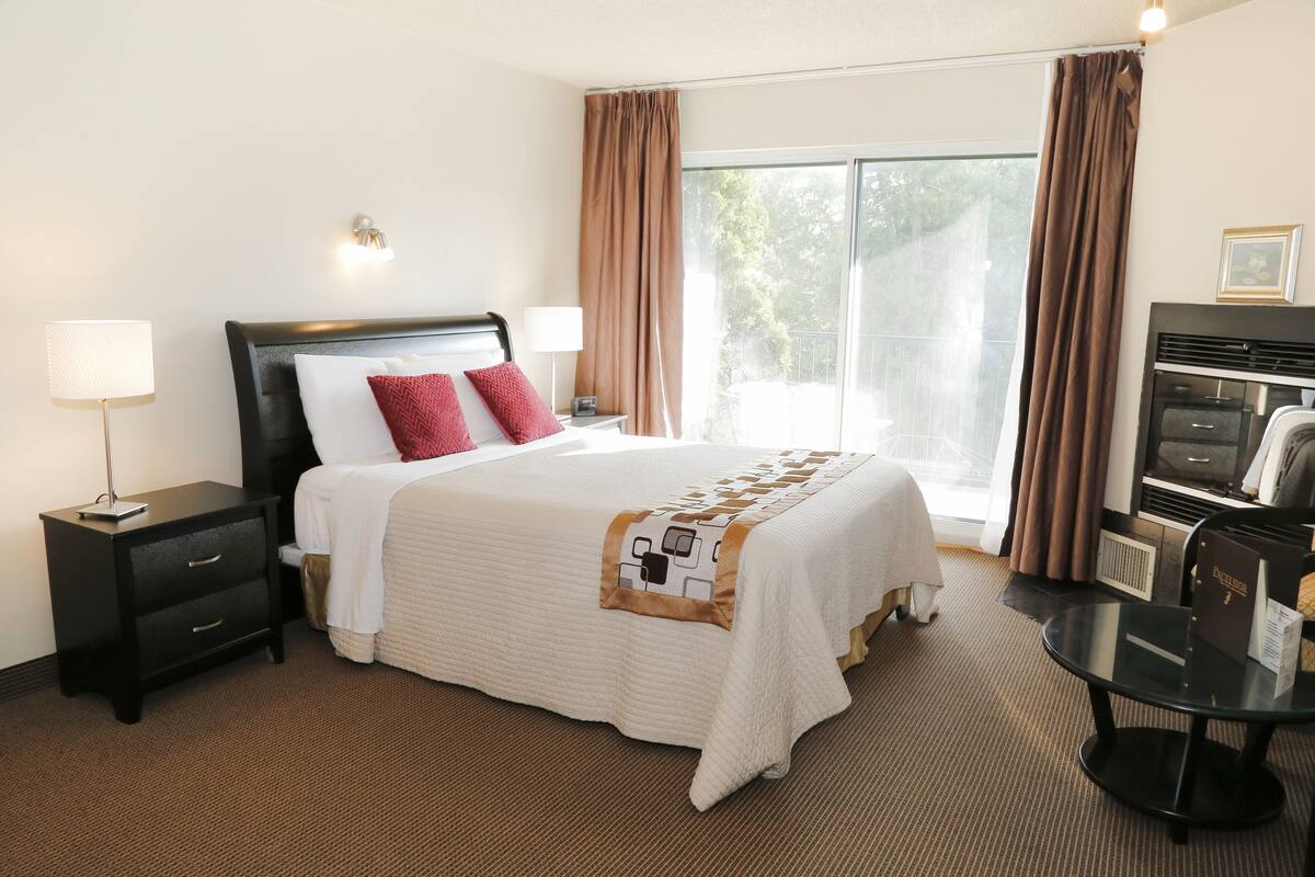 A hotel room with a large bed, two bedside tables, a desk with a chair, a window with curtains, and a view of greenery outside.