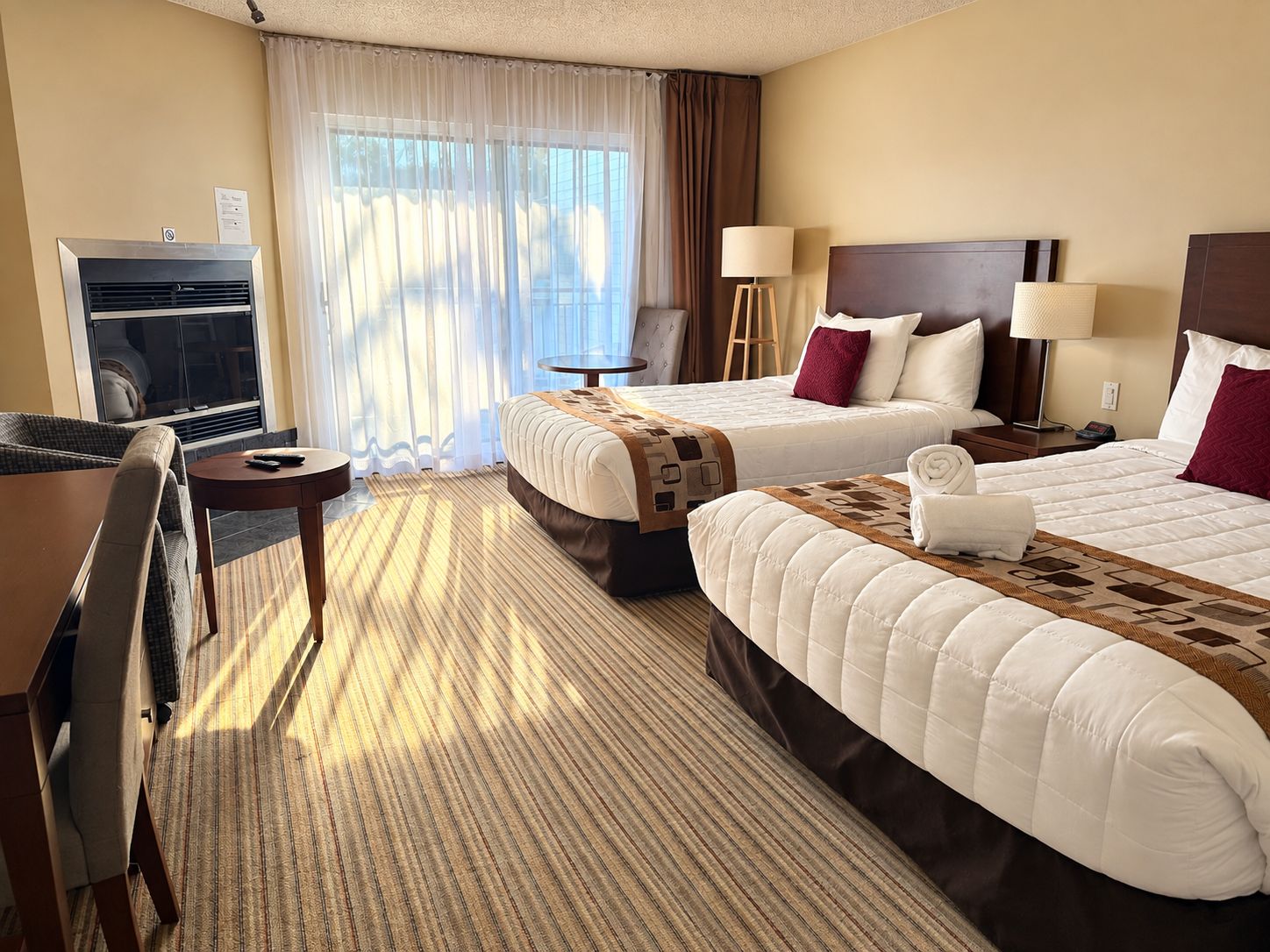 A hotel room with two beds, a desk, a chair, a lamp, and a TV.