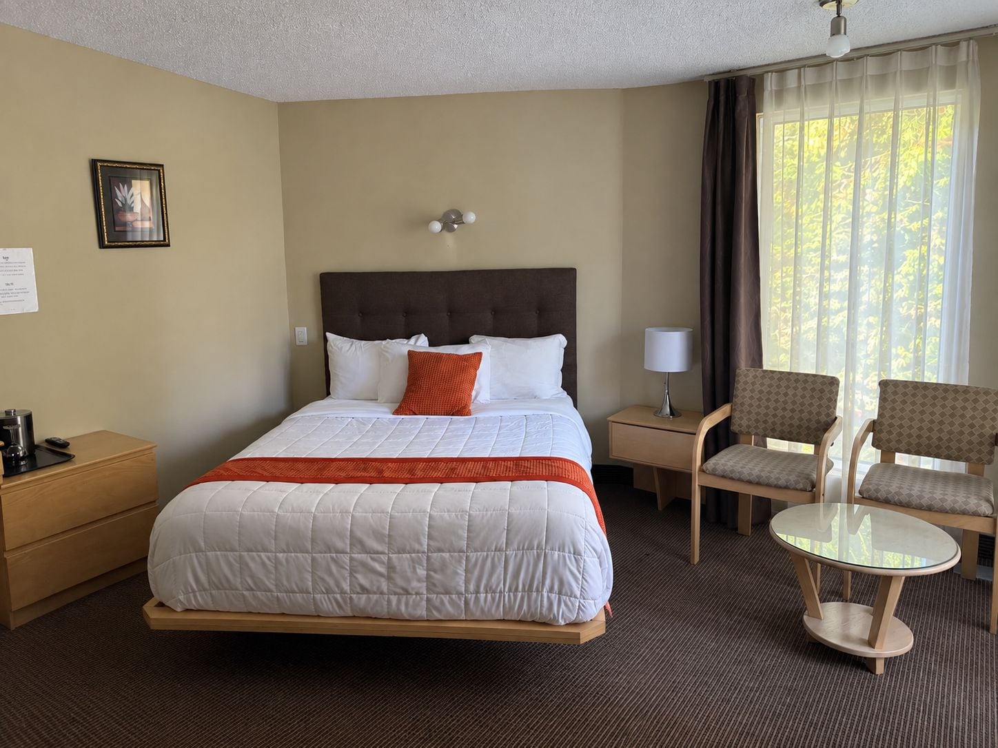 Ein Hotelzimmer mit einem Bett, einem Nachttisch, einem Sessel, einem kleinen Tisch und einem Vorhangfenster.