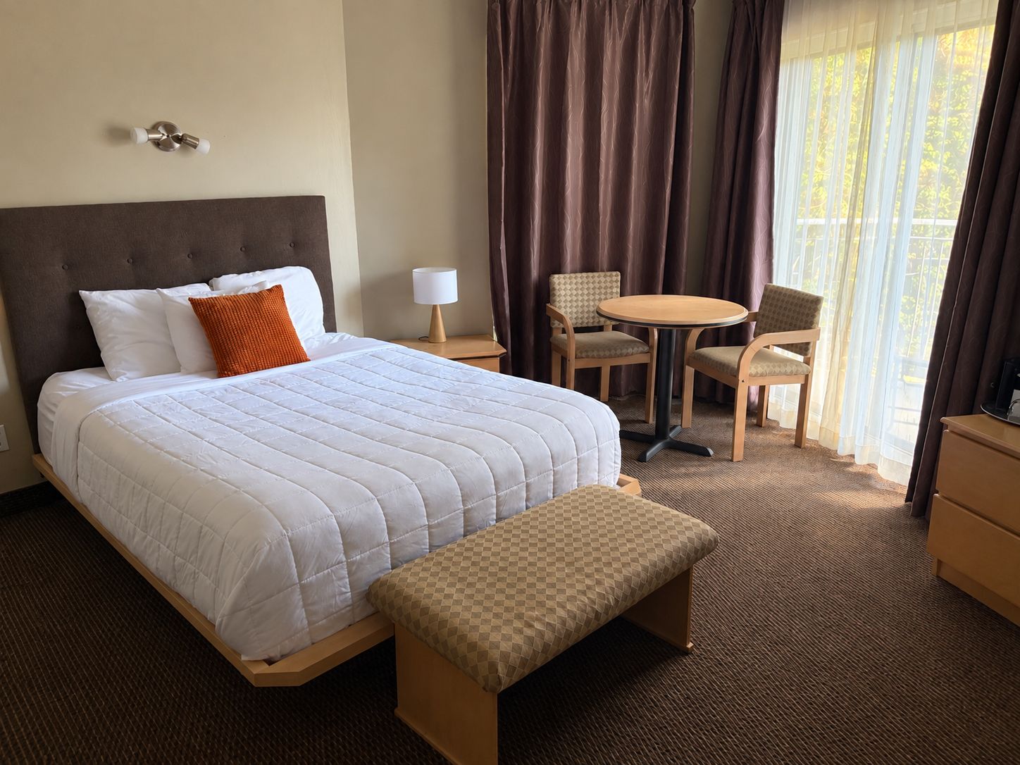 A hotel room with a bed, a bedside table with a lamp, a chair, a small round table, and a bench.