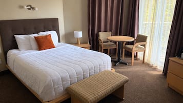 Una habitación de hotel con cama, mesita de noche con lámpara, una silla, una pequeña mesa redonda y un banco.