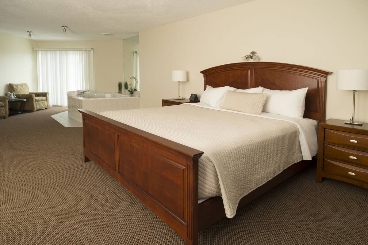 A hotel room with a large bed, a wooden nightstand, a chair, a desk, and a bathroom with a bathtub.