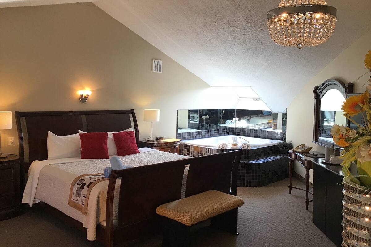 A hotel room with a large bed, a bench, a small table, a mirror, a chandelier, and a view of a bathroom with a bathtub.
