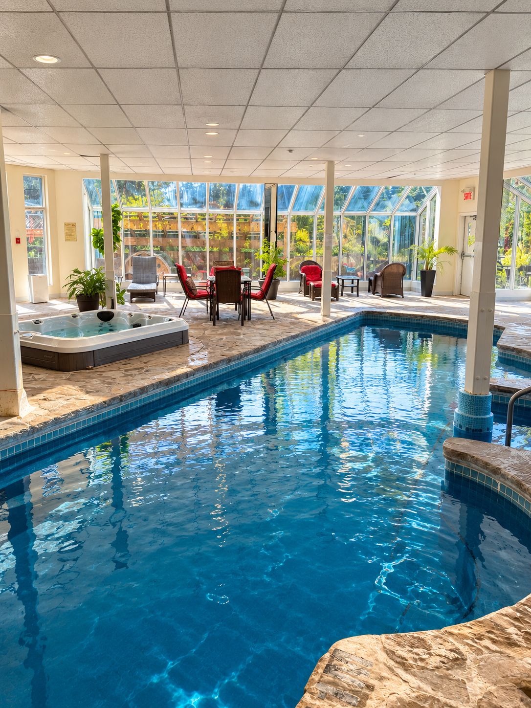 An indoor swimming pool with clear blue water, surrounded by red chairs and tables, and large windows offering a view of greenery outside.